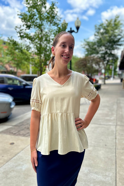 A woman standing outdoors on a sidewalk wearing a cream-colored short-sleeve peplum top with lace sleeve details and a navy blue skirt.