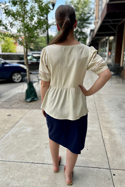 A woman viewed from behind wearing a cream-colored short-sleeve blouse with lace trim on the sleeves and a navy blue skirt, standing on a city sidewalk.