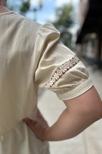 Close-up of a person wearing a cream-colored blouse with short puff sleeves and a floral lace cuff detail, shown from the back in an outdoor setting.
