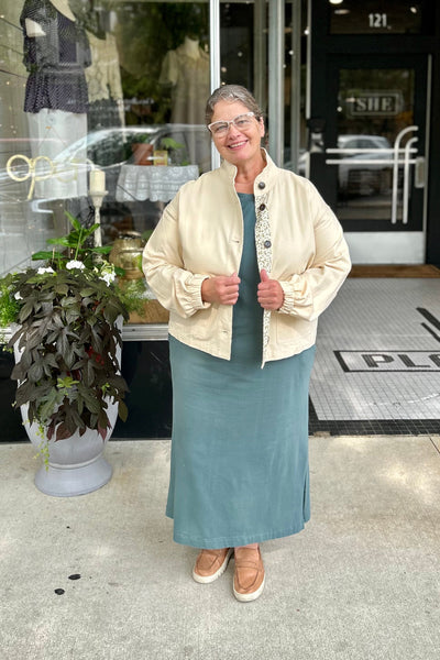 A woman wearing a cream-colored jacket with  detailing on the collar and buttons, paired with a long teal skirt and tan shoes, standing on a sidewalk in front of a boutique storefront.