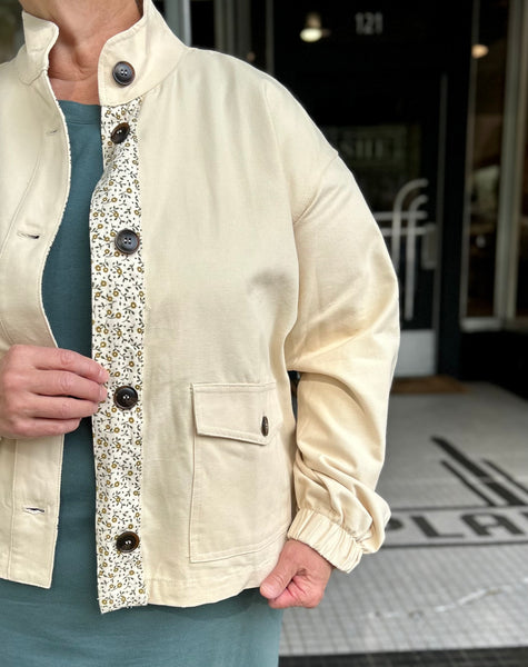 A cream-colored linen jacket with a floral-patterned button placket and dark buttons, worn by a model in front of a storefront.