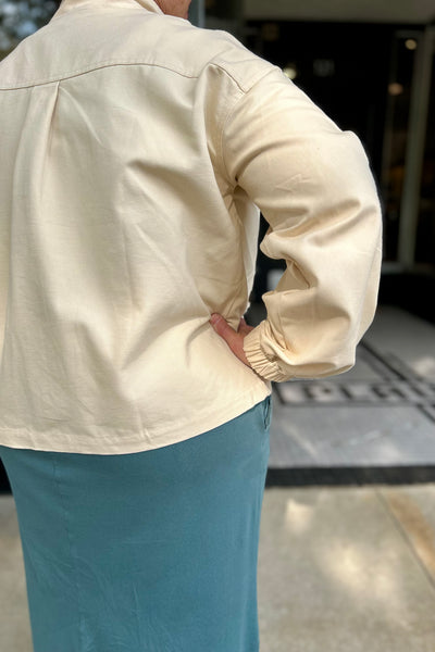 A person shown from the back wearing a cream-colored long-sleeve button-up shirt and a teal skirt, standing outdoors near a building entrance.