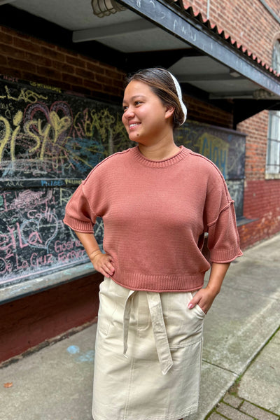 A woman wearing a dusty rose ribbed knit short-sleeve sweater and a beige skirt, standing outdoors in front of a brick building with graffiti.