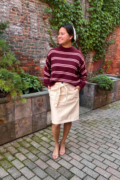A woman stands outdoors in front of a brick wall with green ivy, wearing a burgundy and pink striped knit sweater, a cream corduroy skirt with a tie waist, and clear high-heeled sandals.
