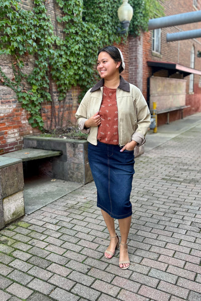 A woman stands on a brick-paved outdoor walkway wearing a beige jacket with dark brown collar, a rust-colored patterned top, and a dark blue midi skirt.