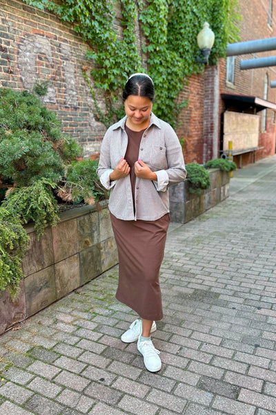 A woman stands on a brick walkway outdoors wearing a light beige button-up shirt over a brown top, a brown midi skirt, and white sneakers.