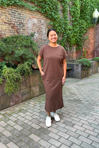 A woman wearing a brown short-sleeve midi dress with side pockets, paired with white sneakers, standing outdoors on a paved walkway in front of a brick wall with green ivy.
