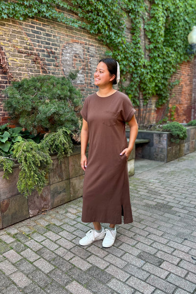 A woman wearing a brown short-sleeve midi dress with side slits and white sneakers, standing outdoors on a paved walkway in front of a brick wall covered in green ivy.