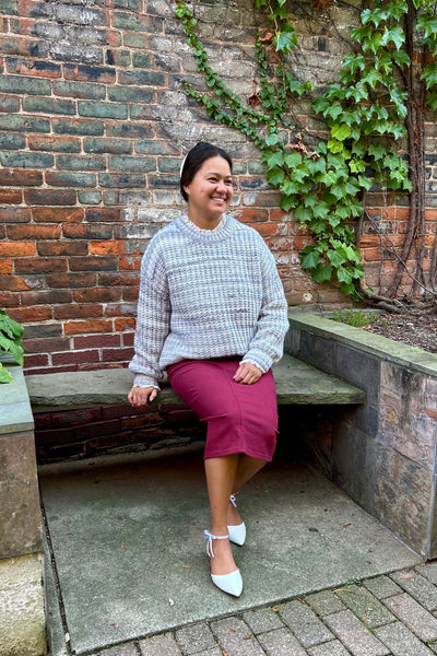 A woman sitting on a stone bench in front of a brick wall with green ivy, wearing a gray and white textured knit sweater, a magenta midi skirt, and white pointed-toe heels.