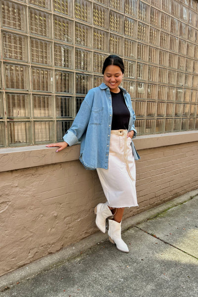 A woman posing outdoors against a glass block wall and concrete sidewalk, wearing a light blue denim shirt over a black top, a cream-colored skirt, and white ankle boots.