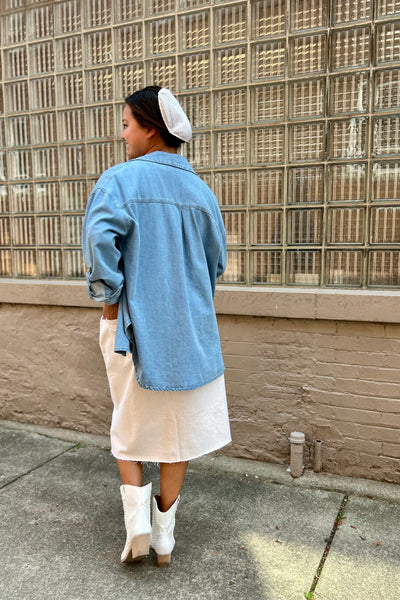 A woman stands outdoors in front of a glass block wall, wearing a light blue denim shirt over a cream dress and white cowboy boots, with a white head covering.