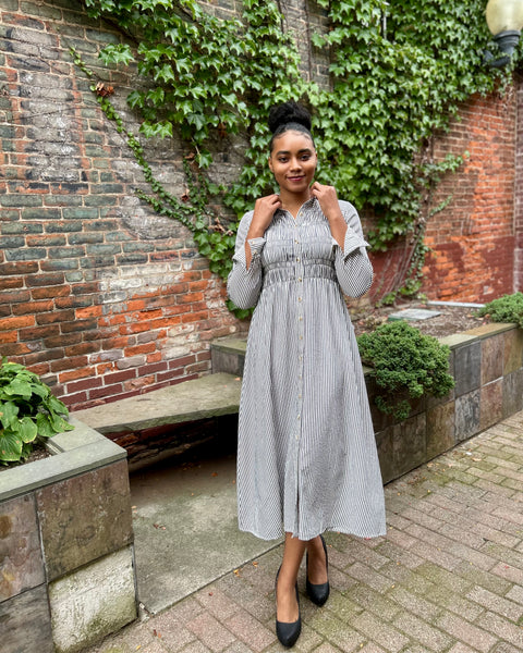 A woman wearing a navy and white vertically striped midi dress with a smocked waist and button-front bodice, standing outdoors in front of a brick wall covered in green ivy.