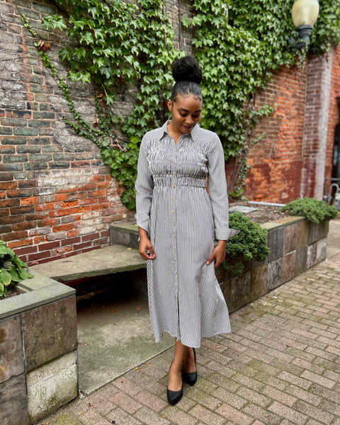 A woman wearing a navy and white vertically striped button-down midi dress with a smocked waist, standing outdoors on a brick walkway in front of a brick wall covered in green ivy.