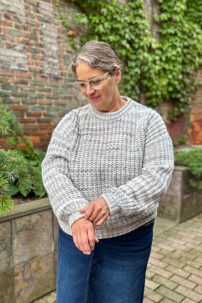 Woman wearing a cozy blue grey variegated sweater with denim skirt standing in front of an ivy covered brick wall. 