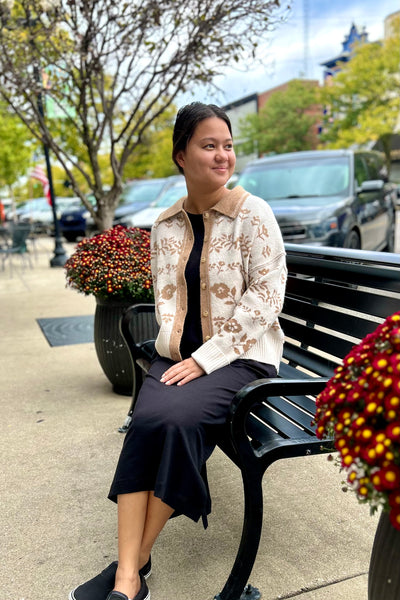 A woman sitting on a black park bench outdoors, wearing a cream and brown floral patterned cardigan over a black top and black midi skirt.