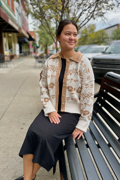 A woman sitting on a black outdoor bench on a city sidewalk, wearing a cream knit cardigan with tan floral patterns and a tan collar, over a black top and black midi skirt.