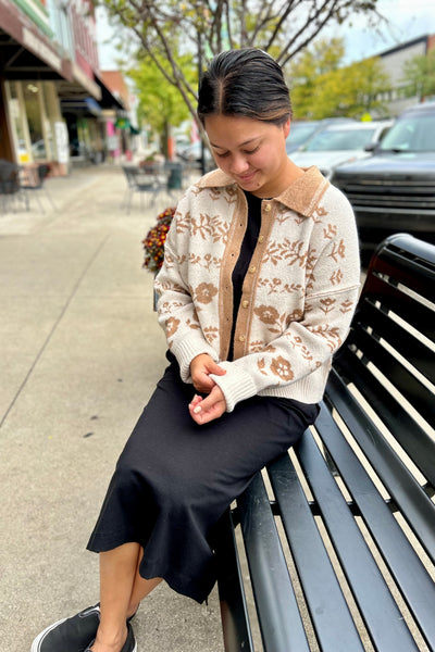 A woman sitting on a black bench outdoors, wearing a cream and tan floral knit cardigan over a black dress .