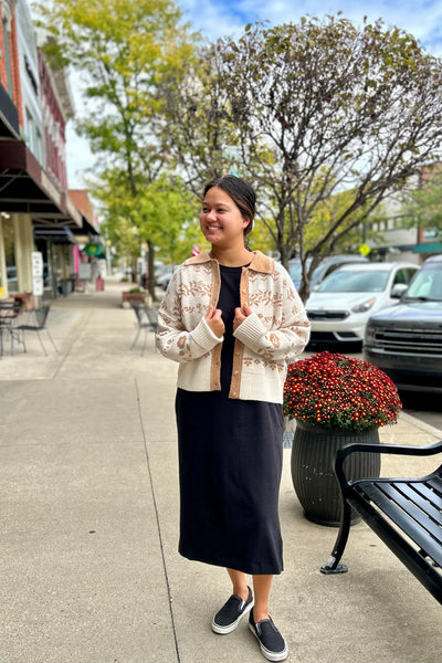 A woman stands on a city sidewalk wearing a cream and tan patterned knit cardigan over a black dress, with black slip-on shoes. The background shows storefronts, parked cars, trees, and a bench.