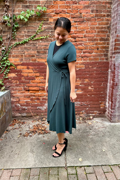 A woman wearing a modest dark teal green faux wrap midi dress with short sleeves and a black V-neck undershirt, standing outdoors against a red brick wall.