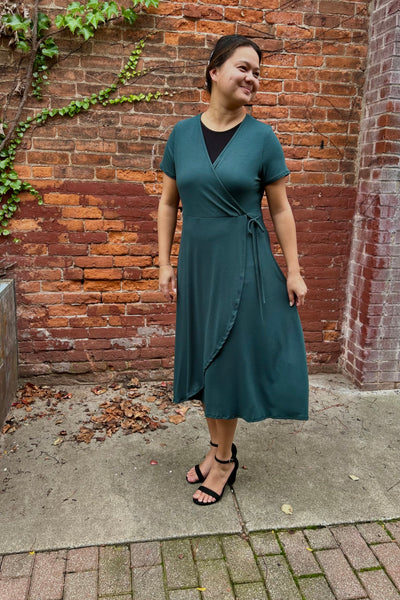 A woman wearing a dark green  wrap midi dress with short sleeves and a side tie, standing outdoors against a red brick wall.
