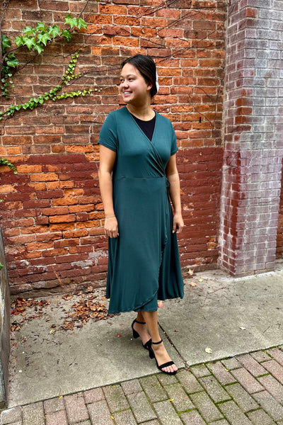 A woman wearing a modest dark green wrap midi dress with short sleeves and a black undershirt, standing on a brick sidewalk in front of a red brick wall.