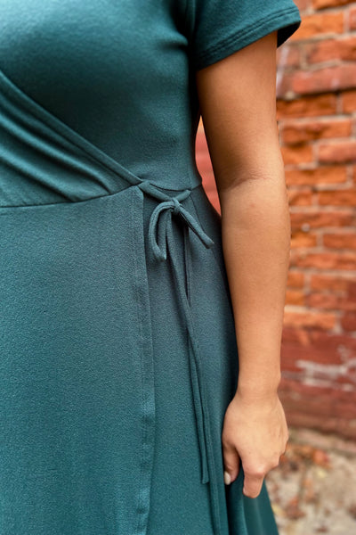A close-up of a person wearing a dark green wrap-style dress with a side tie, standing outdoors against a blurred red brick wall.