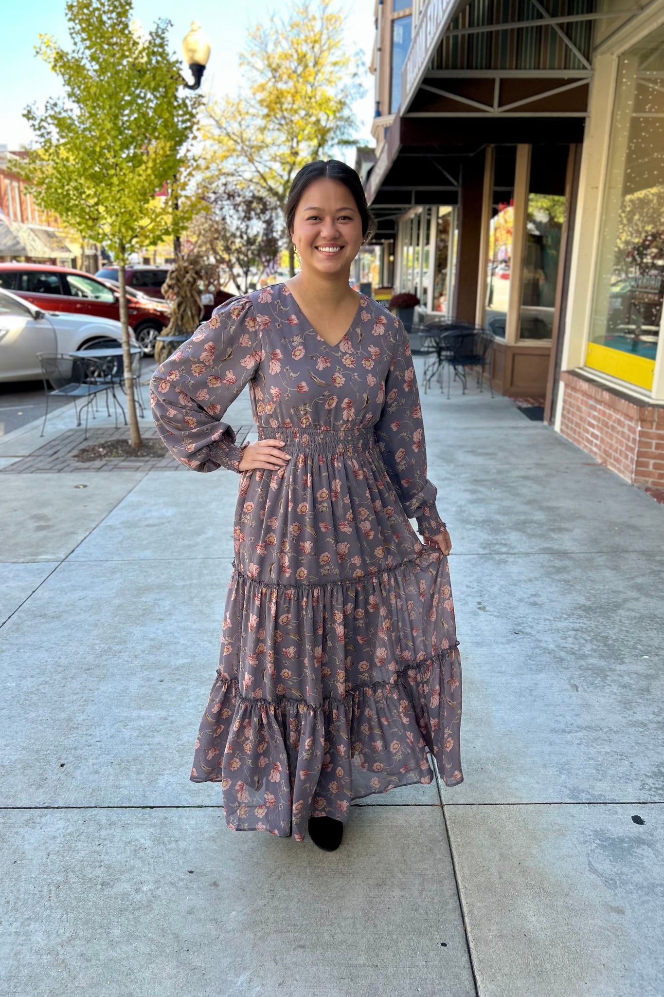 Woman standing on a downtown sidewalk wearing a long floral maxi dress with long sleeves, a V-neckline, and a tiered skirt. The dress has a smocked waist and cuffs, creating a soft, flowy silhouette. She is smiling and holding a portion of the dress hem, with fall trees and shopfronts in the background.