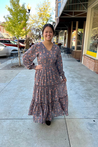 Woman standing on a downtown sidewalk wearing a long floral maxi dress with long sleeves, a V-neckline, and a tiered skirt. The dress has a smocked waist and cuffs, creating a soft, flowy silhouette. She is smiling and holding a portion of the dress hem, with fall trees and shopfronts in the background.