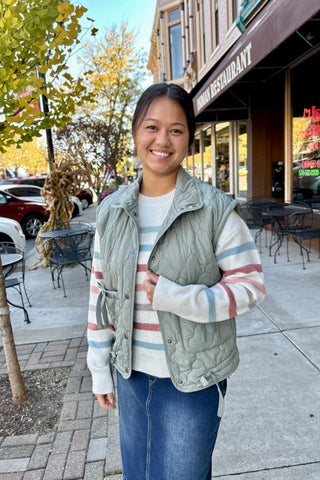 Young woman wearing a sage green quilted puffer vest with a mock neckline and tie-side detail layered over a striped sweater, standing on a fall sidewalk.