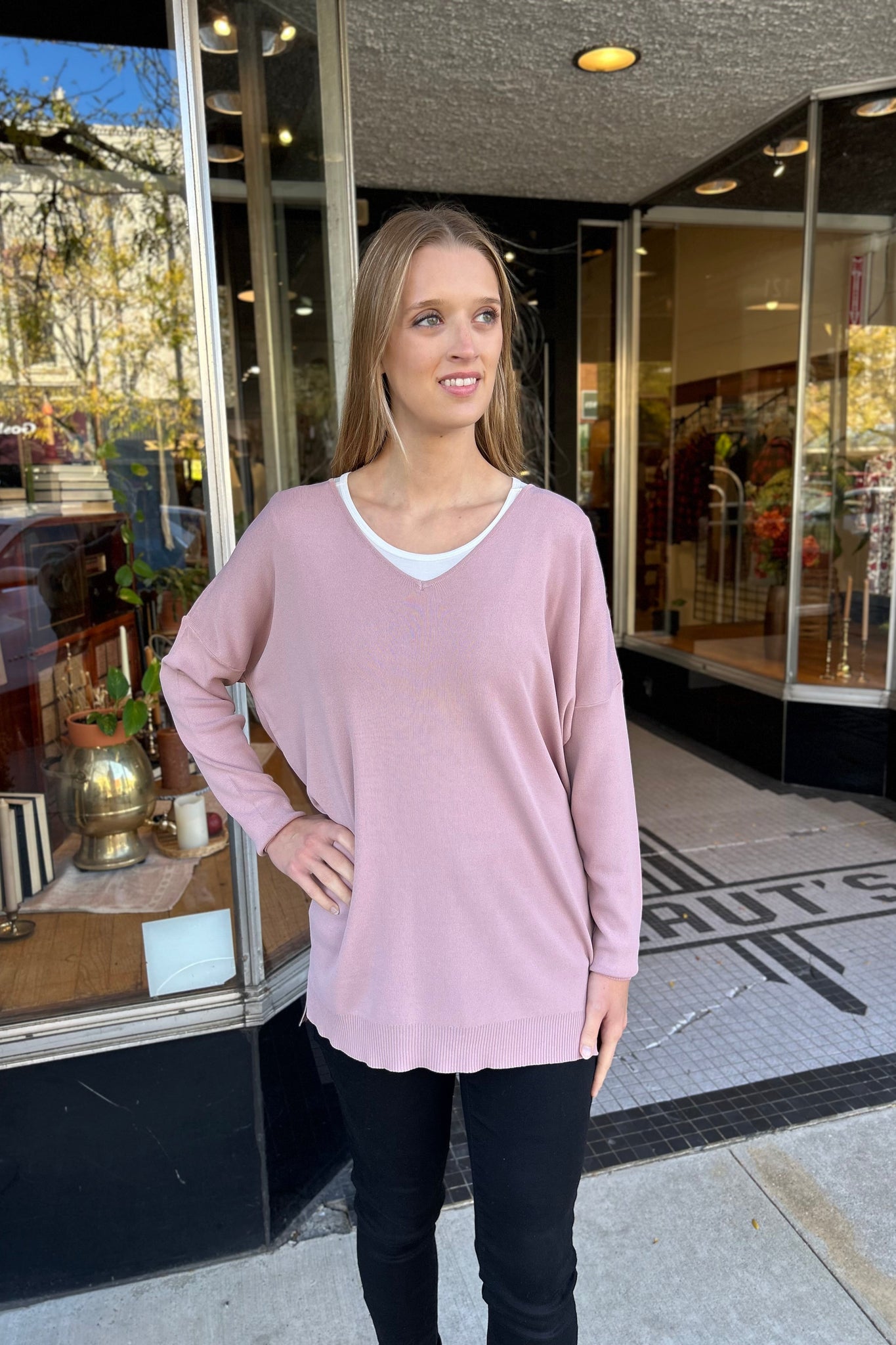 Young woman wearing a dusty mauve V-neck sweater with side slits and high-low hem, paired with black jeans and black boots, standing in front of a boutique shop.