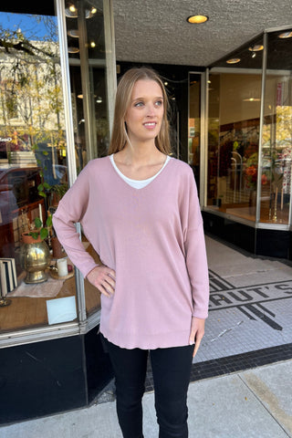 Young woman wearing a dusty mauve V-neck sweater with side slits and high-low hem, paired with black jeans and black boots, standing in front of a boutique shop.