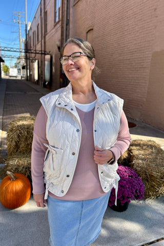 Young woman wearing a quilted puffer vest with a mock neckline and tie-side detail layered over a sweater, standing on a fall sidewalk.