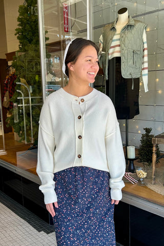 Woman wearing a white cardigan and blue skirt standing in front of a store window display.