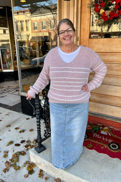 Woman wearing a soft dusty rose striped sweater with a V-neck and relaxed fit, styled with a light denim skirt standing outside a boutique.