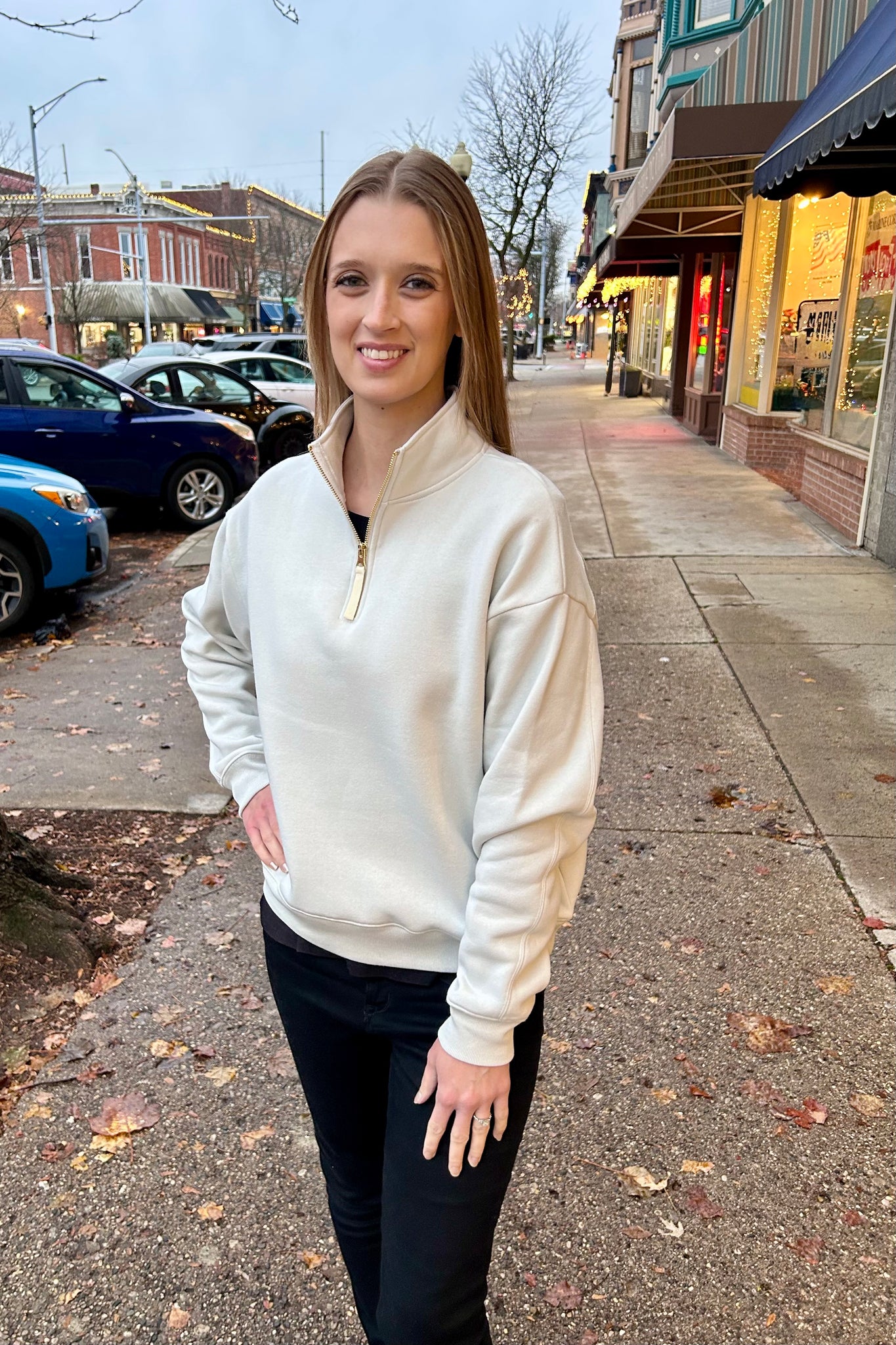 Woman wearing a bone-colored fleece half-zip sweatshirt with gold zipper, standing on a downtown sidewalk with winter lights in the background.