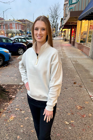 Woman wearing a bone-colored fleece half-zip sweatshirt with gold zipper, standing on a downtown sidewalk with winter lights in the background.