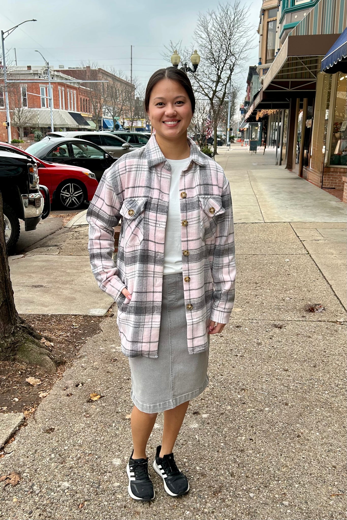Woman wearing a pink and grey plaid flannel shacket with front pockets, paired with a grey skirt on a downtown sidewalk.
