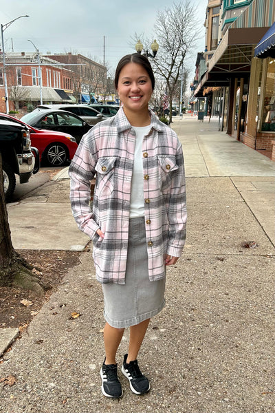 Woman wearing a pink and grey plaid flannel shacket with front pockets, paired with a grey skirt on a downtown sidewalk.