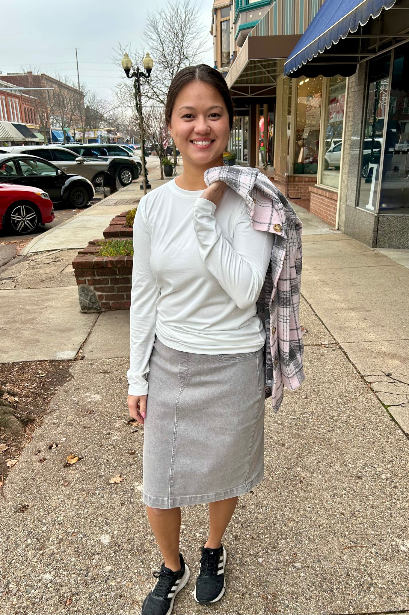 Woman wearing a simple white long sleeve top and grey skirt standing on a downtown sidewalk.