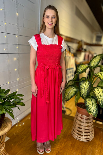 Woman wearing a fuchsia dress with a white shirt underneath, standing indoors with plants and a blurred background.