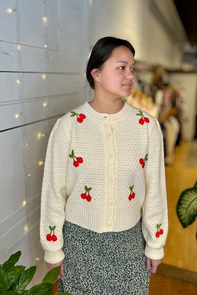 Woman wearing a cream-colored cardigan with cherry designs in an indoor setting.