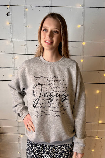 Woman wearing grey Jesus Sweatshirt standing in front of a lighted wall. 