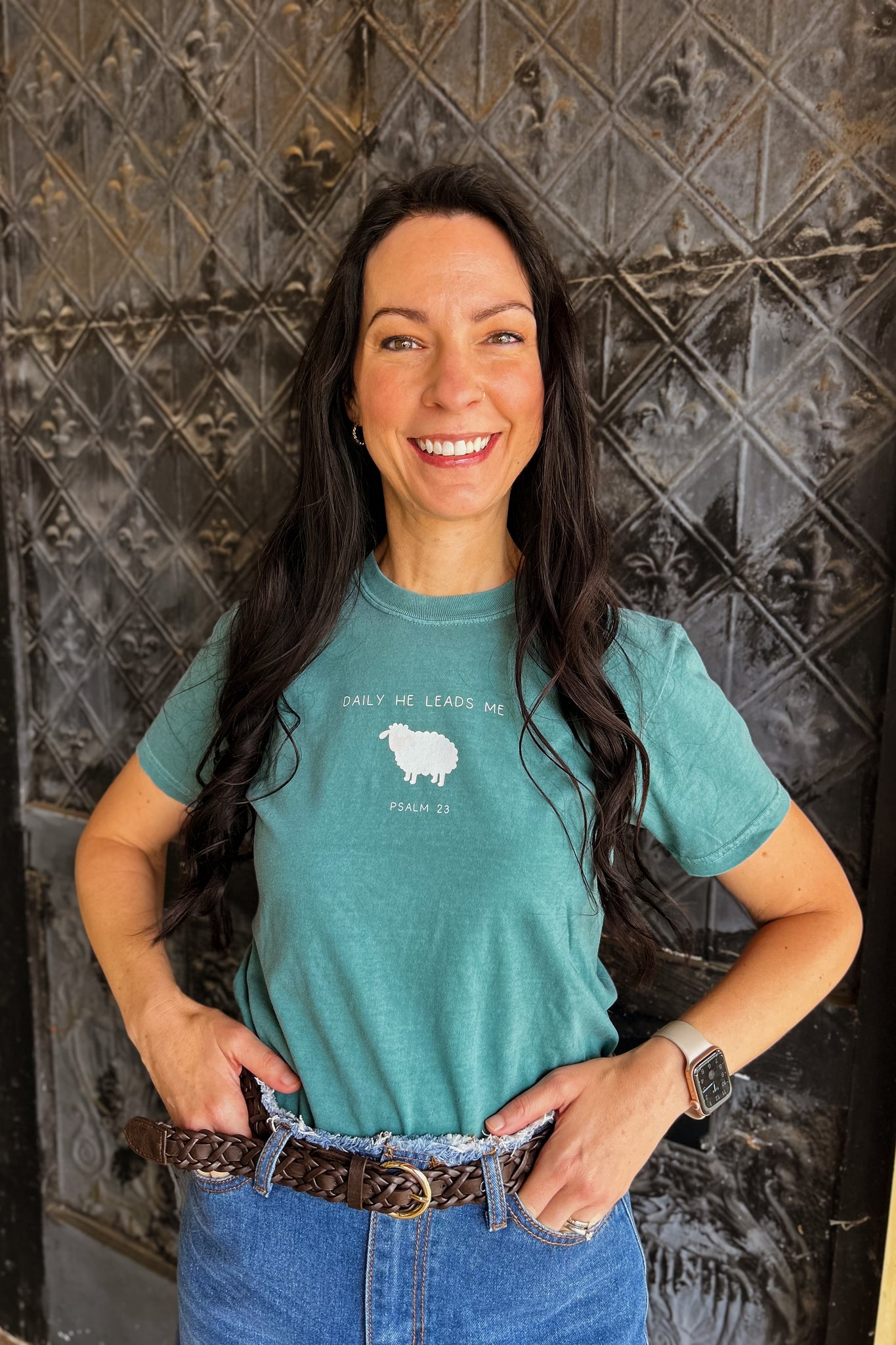 Woman wearing earthy green graphic tee with “Daily He leads me” design featuring a sheep, crew neck, and relaxed fit, styled casually.