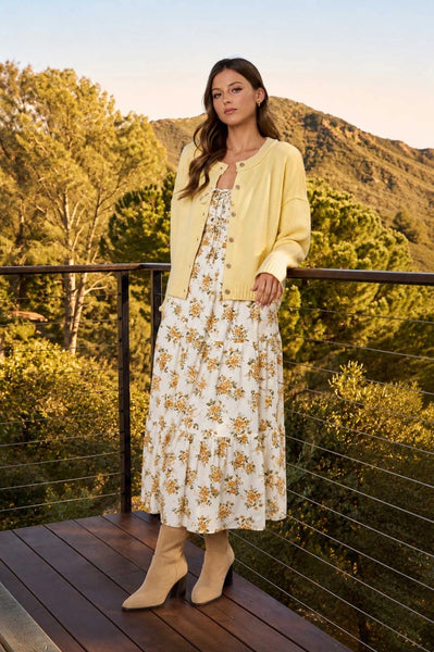 Woman in a floral dress and yellow cardigan standing on a wooden deck with a mountainous background.