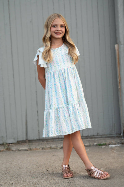 Young girl wearing a light blue floral dress standing against a gray wall.
