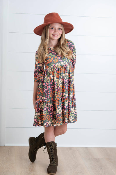 Little girl wearing a floral tiered dress with three-quarter sleeves in warm autumn colors, paired with a rust wide-brim hat for a classic modern modest look.