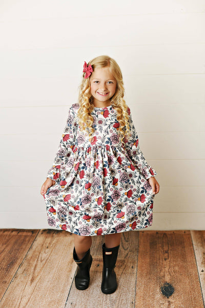 Young girl wearing a floral dress standing on a wooden floor with a white wall background