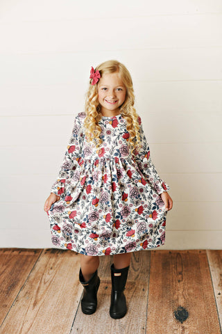 Young girl wearing a floral dress standing on a wooden floor with a white wall background