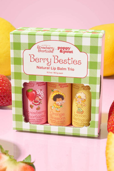 A boxed lip balm trio called Berry Besties by poppy & pout, shown in a green-and-white gingham box with three pastel lip balm tubes visible. The box is surrounded by fresh strawberries, sliced lemons, and orange slices on a pink background.