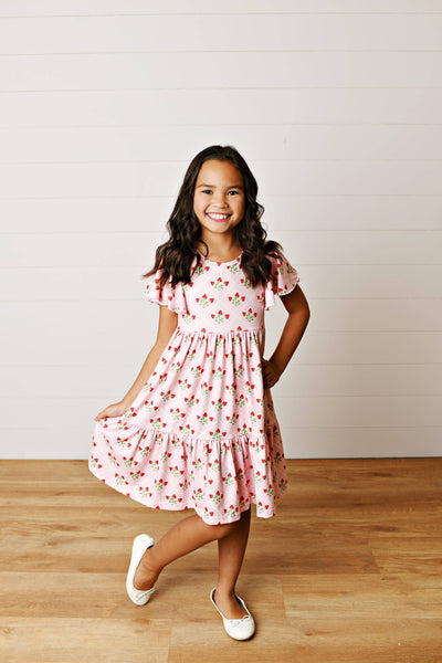 Little girl wearing a pink strawberry print dress with flutter sleeves and a tiered twirl skirt, smiling in a playful modern modest style.